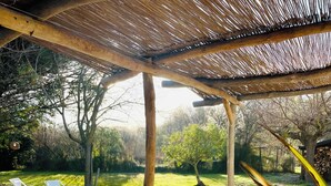 Un área de estar al aire libre con techo, dos sillones blancos, una pérgola de madera y un jardín con árboles y césped.