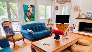 Olohuoneessa on sininen sohva, puinen sohvapöytä, punottuista materiaaleista valmistetut jalkarahit, televisio ja takka.