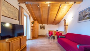 A wooden ceiling with exposed beams, a flat-screen TV on a wooden cabinet, a red sofa, and a dining area with a table and chairs. - Apartments Agave 169A (Ortisei)