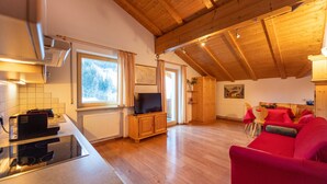 A spacious living area with a wooden ceiling, a red sofa, a dining table with chairs, and a kitchen area with a stove and a television. - Apartments Agave 169A (Ortisei)