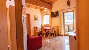 A cozy room with wooden floors, a red sofa, a wooden dining table with chairs, a TV mounted on the wall, and a window with curtains. - Apartments Agave 169B (Ortisei)
