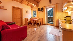 A cozy living room with a red sofa, wooden dining table, and chairs. A wooden door and a TV mounted on the wall are also visible. - Apartments Agave 169B (Ortisei)