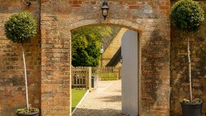 A brick archway with a white door, flanked by potted plants, leading to a grassy area with a wooden fence. - The Hayloft - sleeps 6 guests  in 3 bedrooms (Winchcombe)