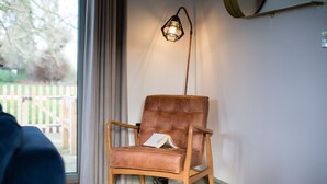 En brun läderfåtölj med en bok, placerad nära ett fönster med gardiner, och en väggmonterad lampa.