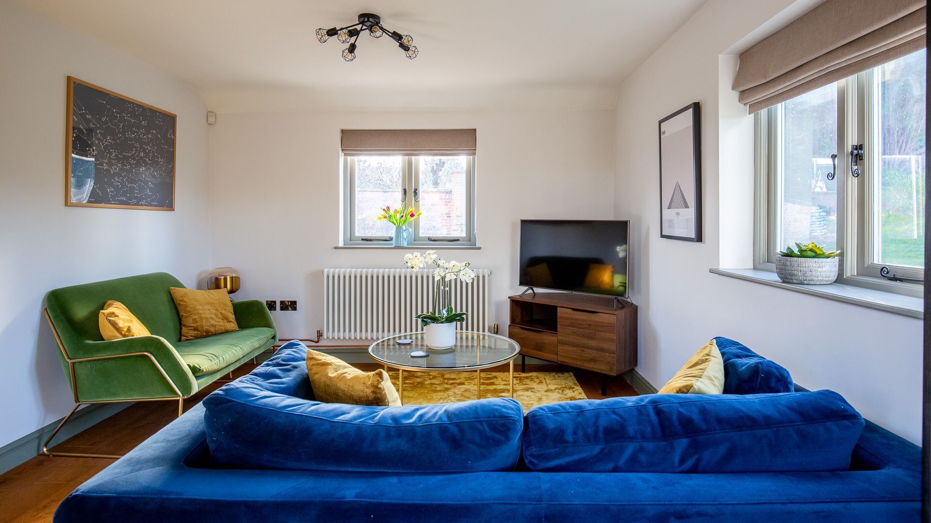 A living room with a blue sofa, a green armchair, a glass coffee table, a TV, and a radiator.