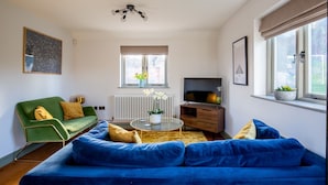 Ett vardagsrum med en blå soffa, en grön fåtölj, ett soffbord i glas, en tv och ett element.