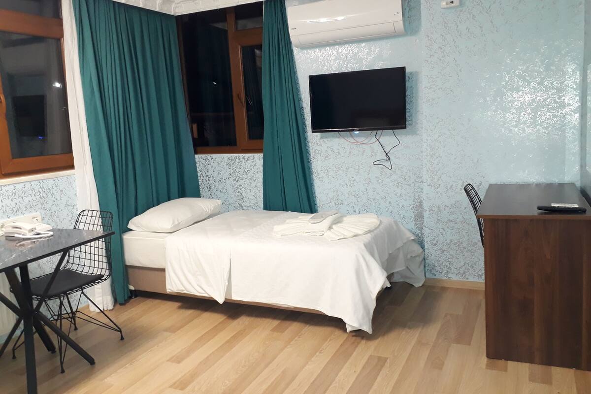 A room with a bed, a desk with a chair, a TV mounted on the wall, and an air conditioning unit.