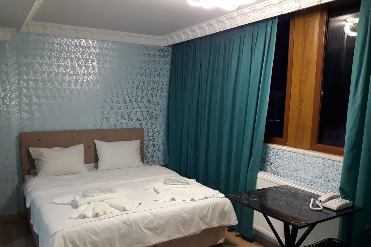 A bedroom with a bed, a table with a phone, and a window with curtains.