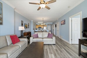 A living room with a beige sofa, a red pillow, a coffee table, and a ceiling fan. - Beautiful Beachfront Condo with Amazing Views and Beachfront Pool! Chair Service Included* (Panama City Beach)
