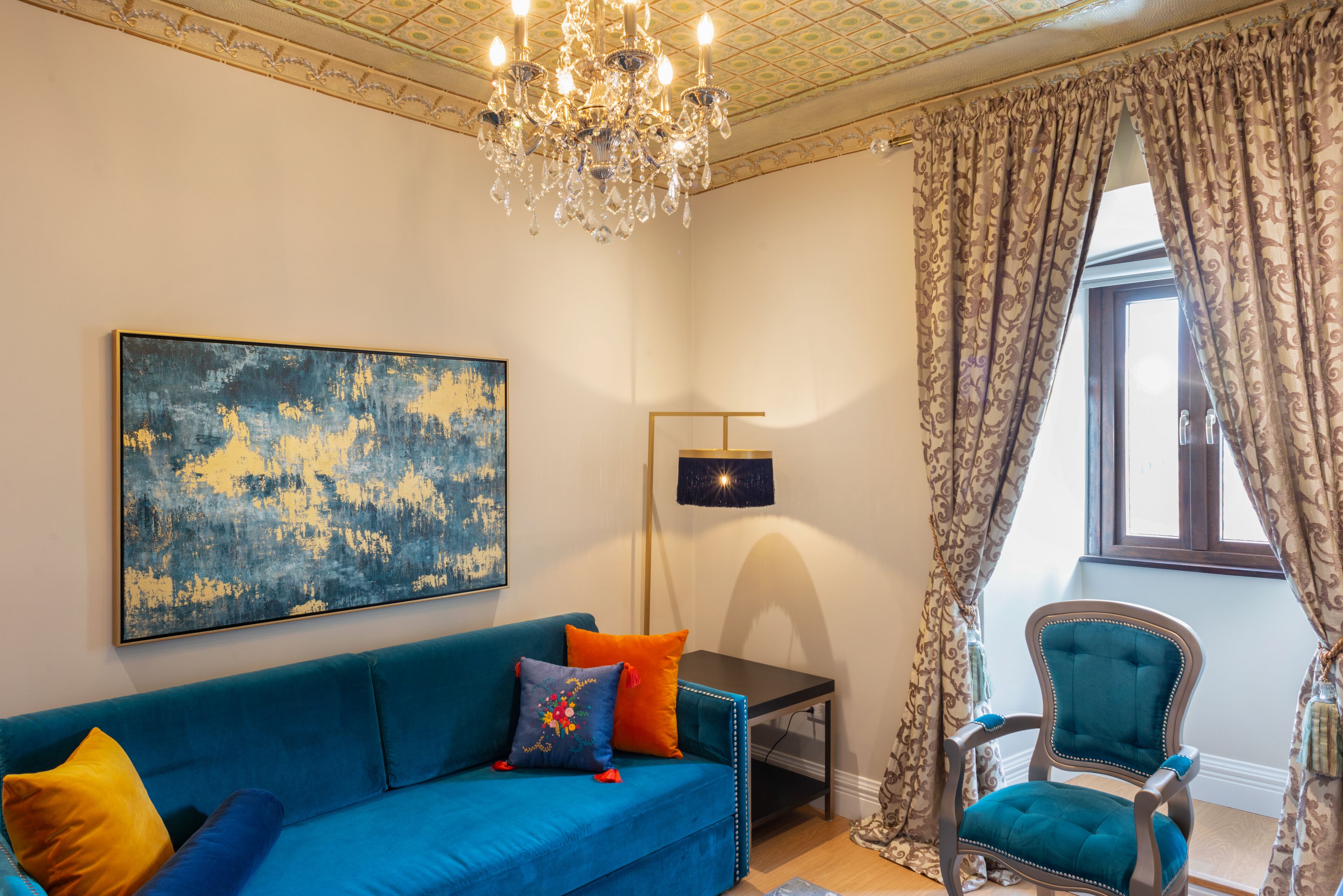 Una habitación con un sofá azul, una silla decorativa, una araña y un cuadro abstracto grande.
