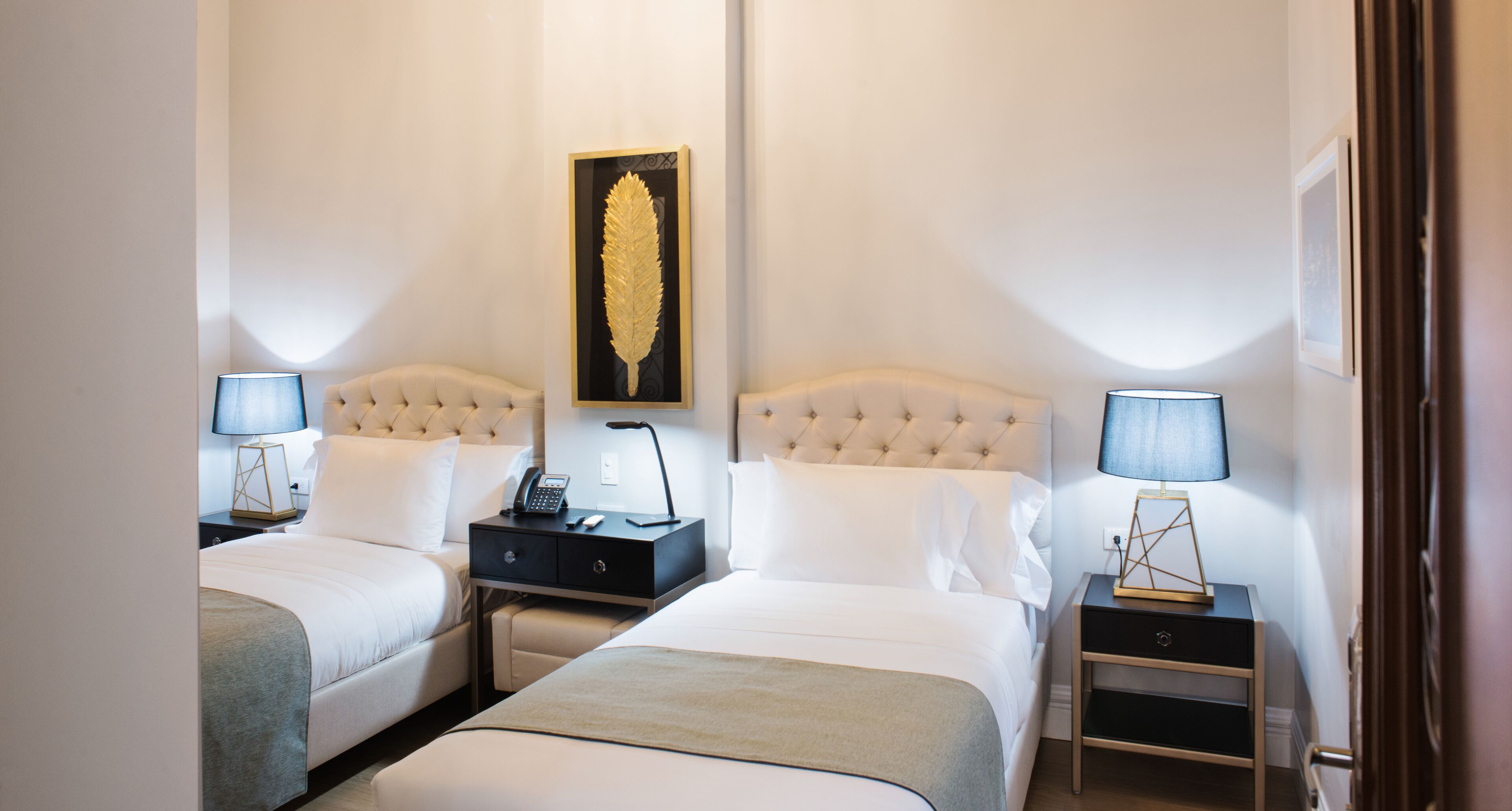 A hotel room with two beds, a nightstand with a lamp, a framed artwork on the wall, and a small table with a lamp.