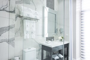 Un baño moderno con paredes de mármol, cabina de ducha de vidrio, lavabo blanco y tocador con espejo.