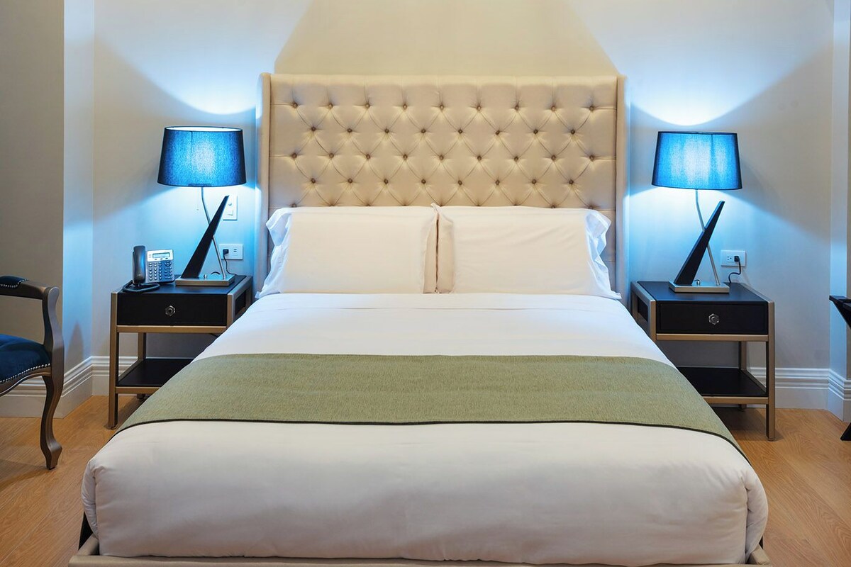 A bedroom with a tufted headboard, two bedside tables with lamps, a chandelier, and a green throw blanket.
