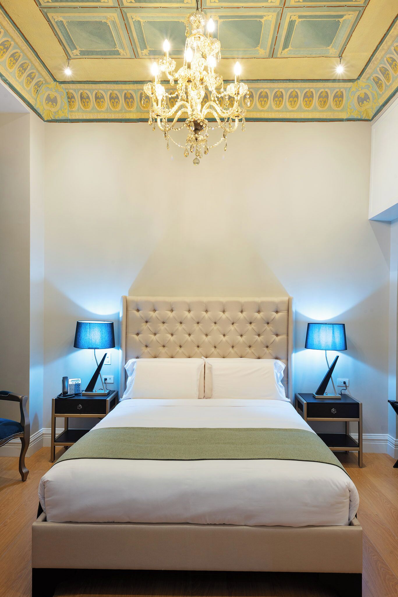 Une chambre à coucher avec une tête de lit capitonnée, deux tables de chevet avec des lampes, un lustre et un plaid vert.