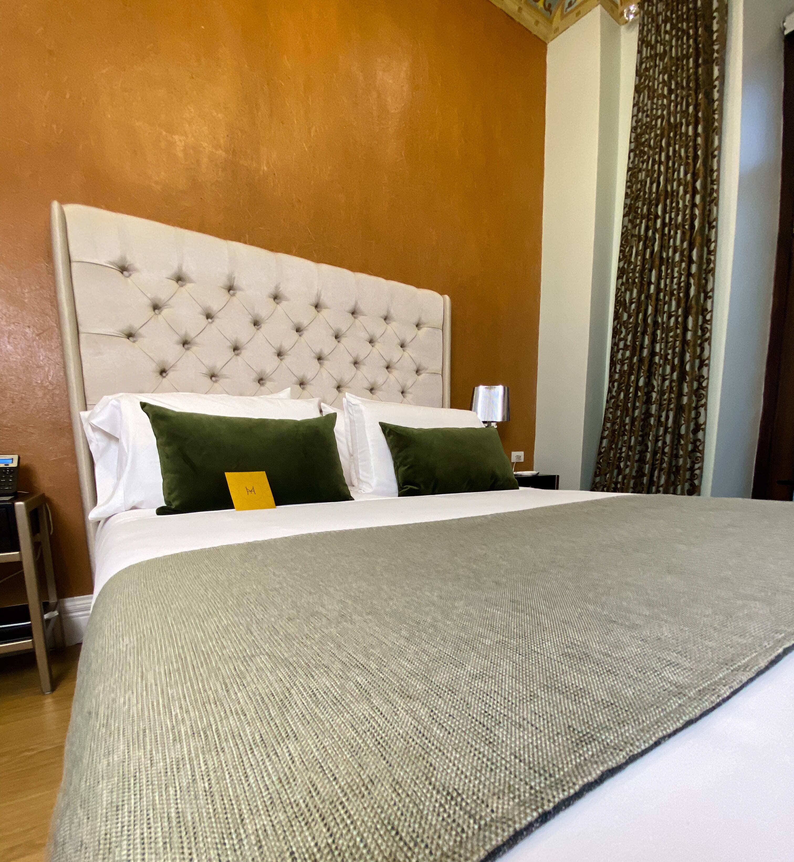 Ein Hotelzimmer mit einem gepolsterten Kopfteil, einem Bett mit weißen und grünen Kissen, einer grauen Tagesdecke und einem kleinen Nachttisch mit einer Lampe.