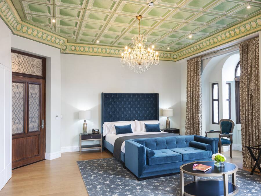 A bedroom with a large bed, a blue sofa, a chandelier, and a view of the outside.