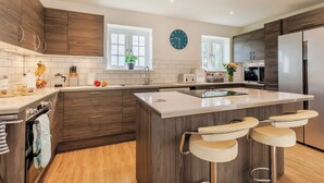 A modern kitchen with wooden cabinets, a large island with bar stools, a sink, and a clock on the wall. - 1 bedroom accommodation in Ocle Pychard (Ocle Pychard)