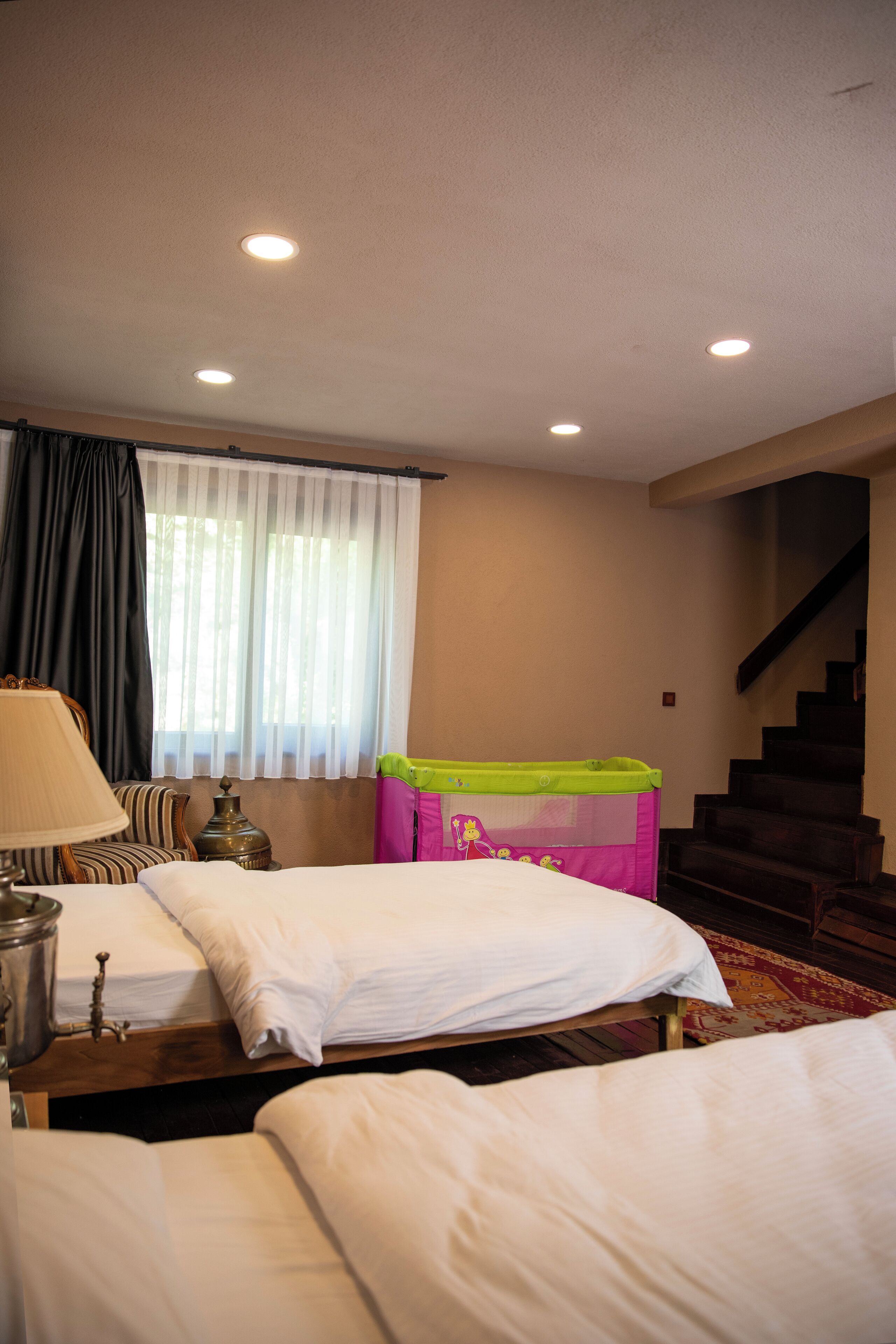 A hotel room with two beds, a window with sheer curtains, a lamp, a radiator, and a staircase.