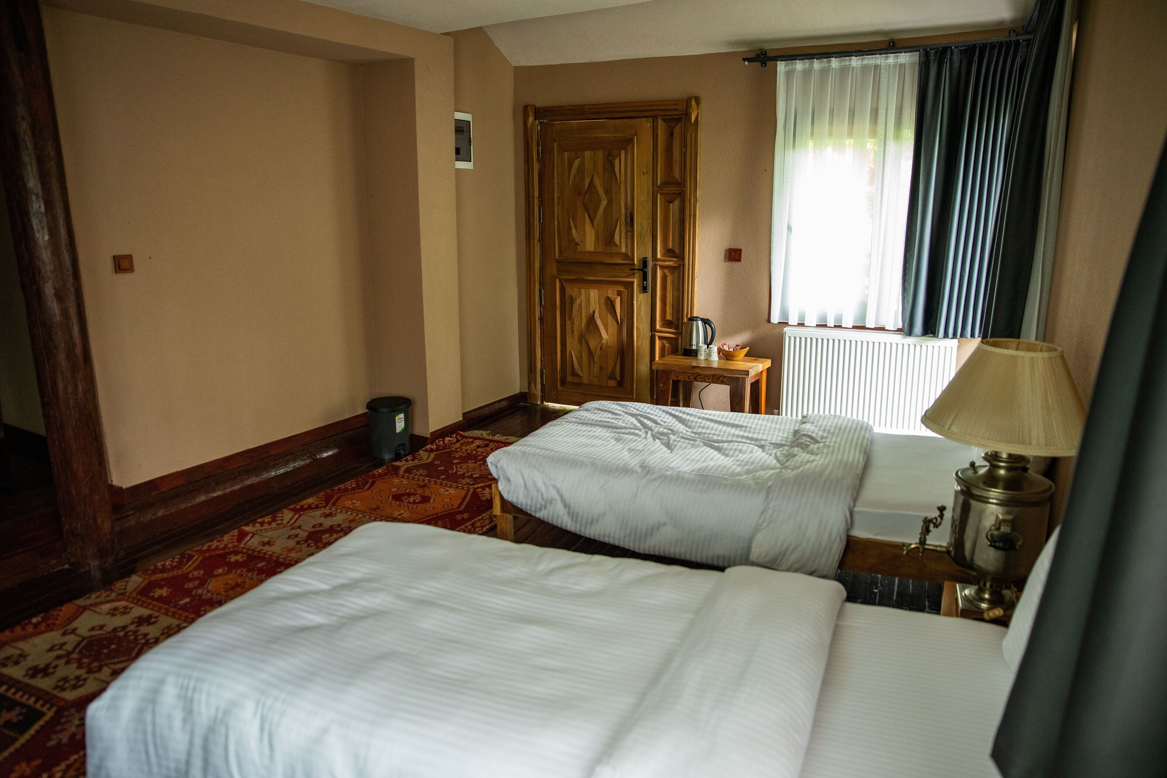 A hotel room with two beds, a wooden door, a radiator, a table with a lamp, and a window with curtains.