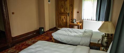 A hotel room with two beds, a wooden door, a radiator, a table with a lamp, and a window with curtains.