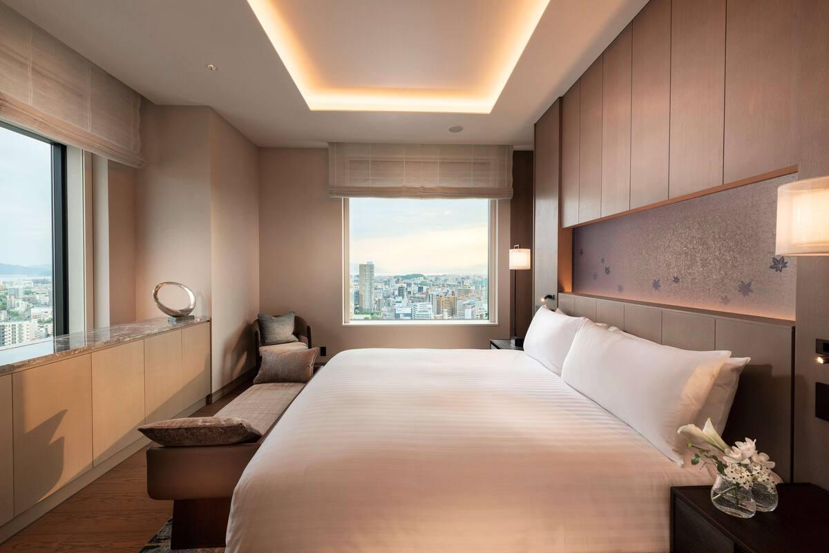 Una habitación de hotel moderna con una cama grande, vistas a la ciudad, una mesita de noche con lámpara y una ventana con panorama urbano.
