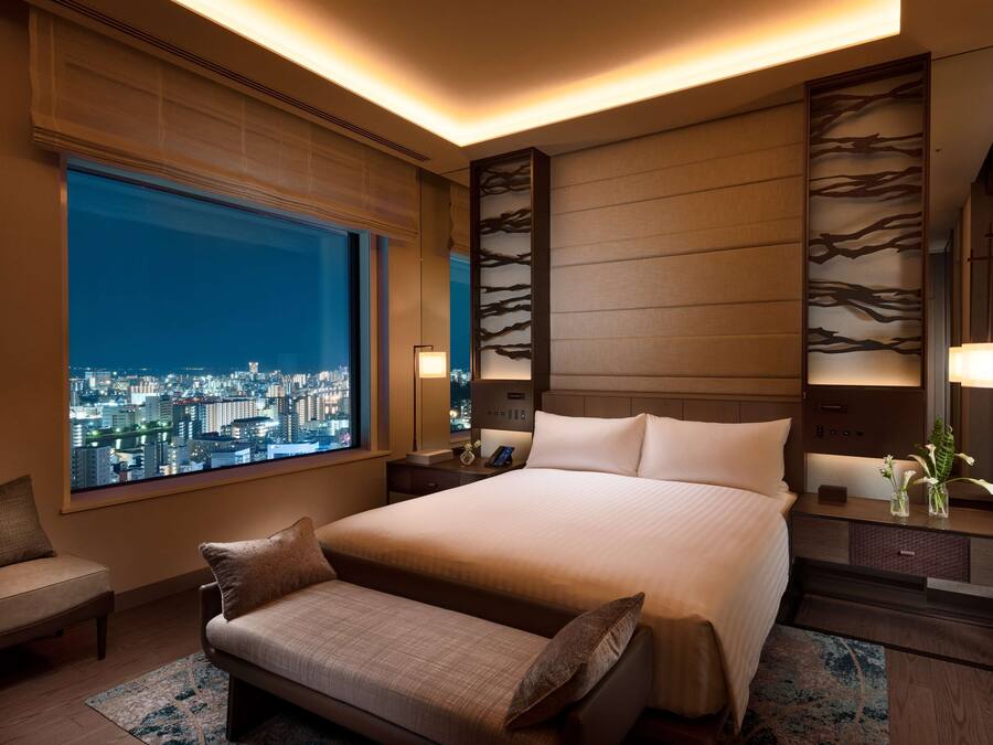 Una habitación de hotel moderna con una cama grande, mesitas de noche, un banco, un ventanal con vistas a la ciudad y un panel decorativo en la pared.