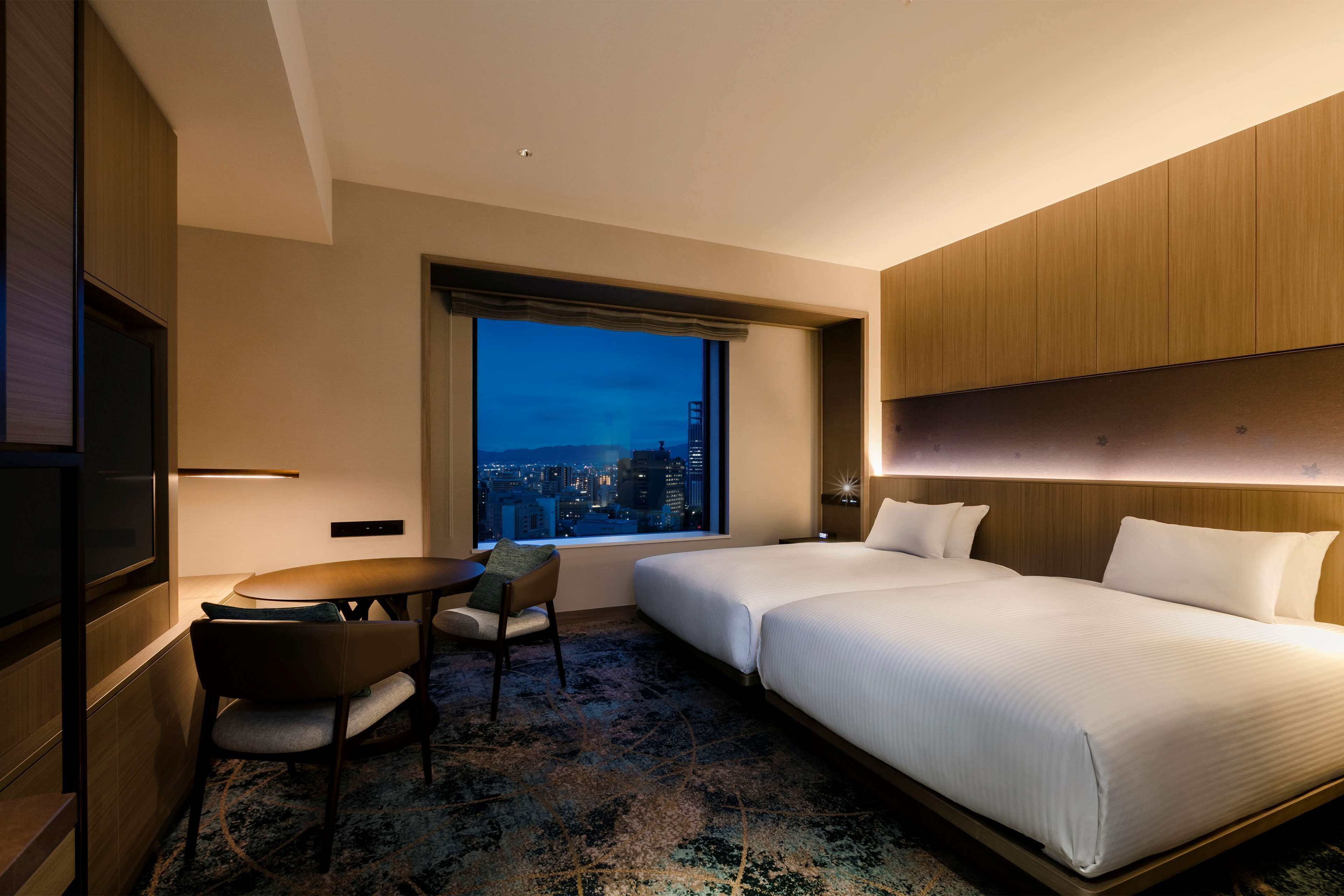 Ein Hotelzimmer mit zwei Betten, einem Schreibtisch, einem Stuhl und einem großen Fenster mit Blick auf die Stadt.