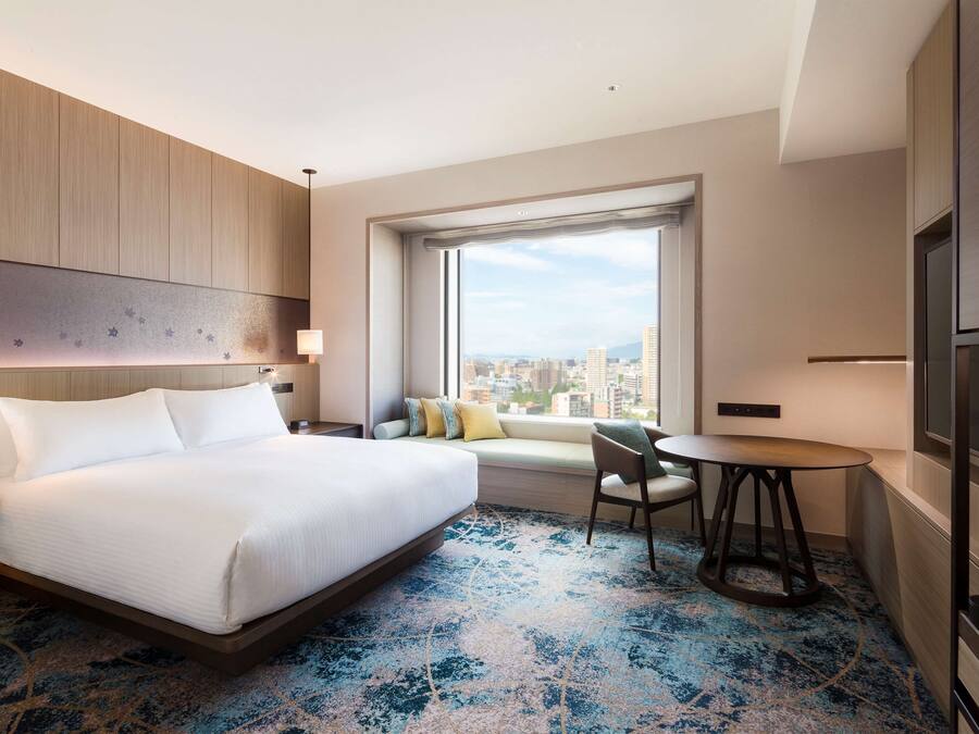 A modern hotel room with a large bed, a seating area by the window, a small table, and a TV.
