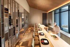 A modern restaurant with a long counter, wooden flooring, and a chef preparing food.