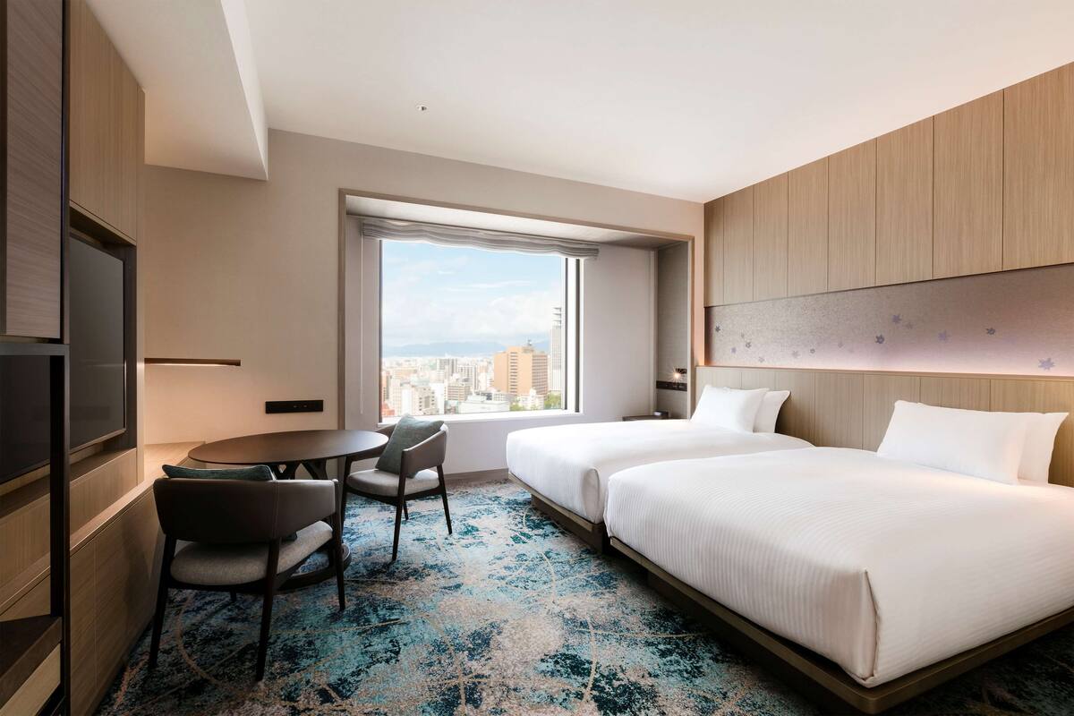 Habitación de hotel con dos camas, un escritorio, una silla y un amplio ventanal con vista a la ciudad.