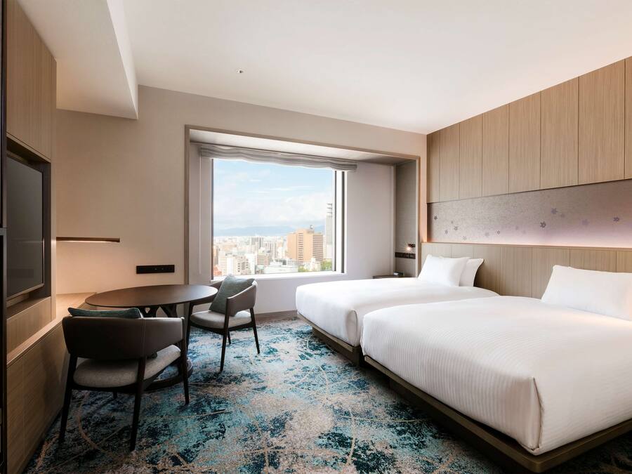Habitación de hotel con dos camas, un escritorio, una silla y un amplio ventanal con vista a la ciudad.