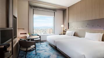 Ein Hotelzimmer mit zwei Betten, einem Fernseher, einem kleinen Tisch mit Stuhl und einem großen Fenster mit Blick auf die Stadt.