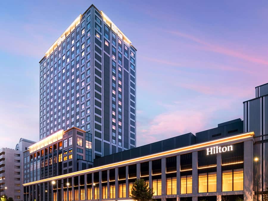 Un moderno edificio de hotel con un llamativo letrero Hilton, que presenta una estructura alta con numerosas ventanas y una fachada bien iluminada.