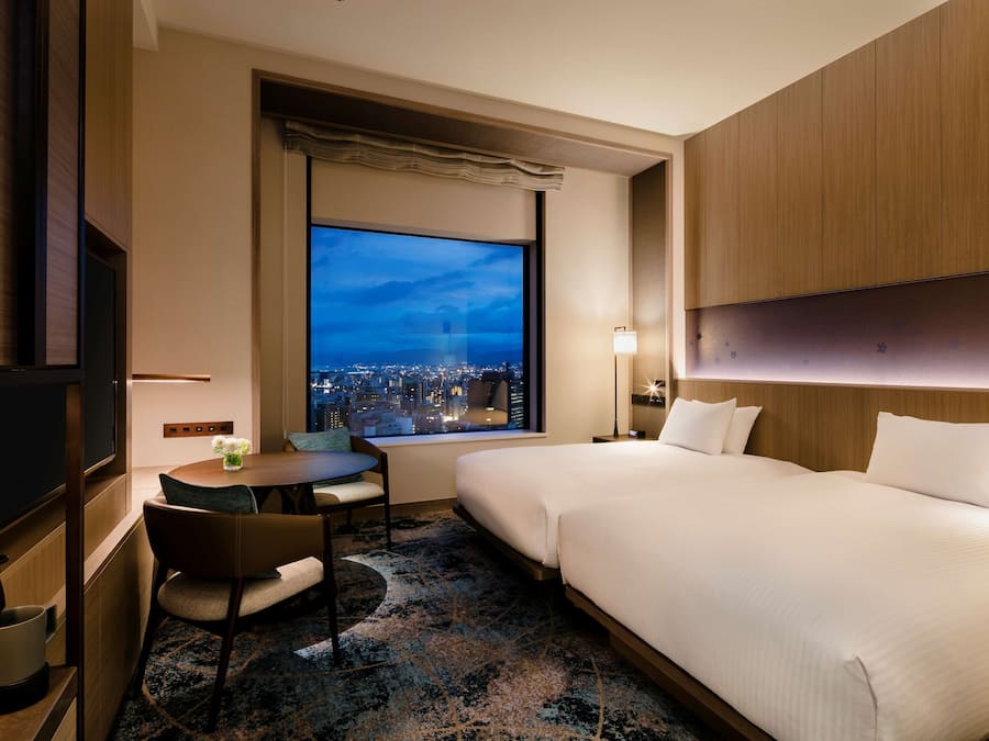 A hotel room with a large window, a bed, a desk with a chair, and a TV.