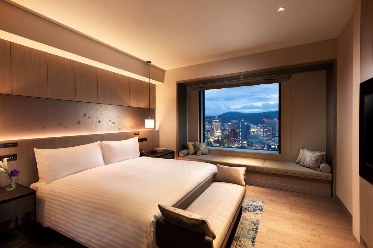 Una habitación de hotel moderna con una cama grande, una zona de estar con vistas a la ciudad y un ventanal amplio.