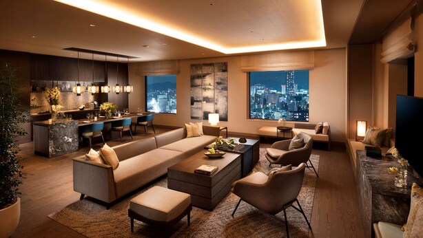 Una sala de estar moderna con sofá, mesa de centro y sillas, un área de cocina con asientos tipo barra y ventanas amplias que ofrecen vistas de la ciudad.