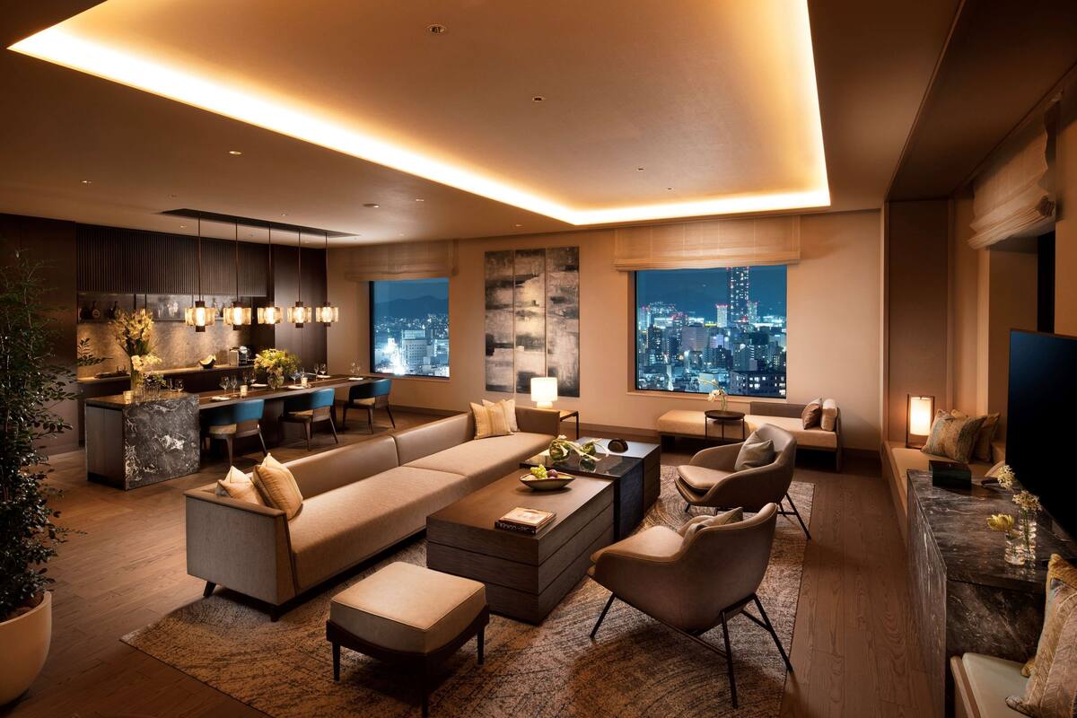 Una sala de estar moderna con sofá, mesa de centro y sillas, un área de cocina con asientos tipo barra y ventanas amplias que ofrecen vistas de la ciudad.