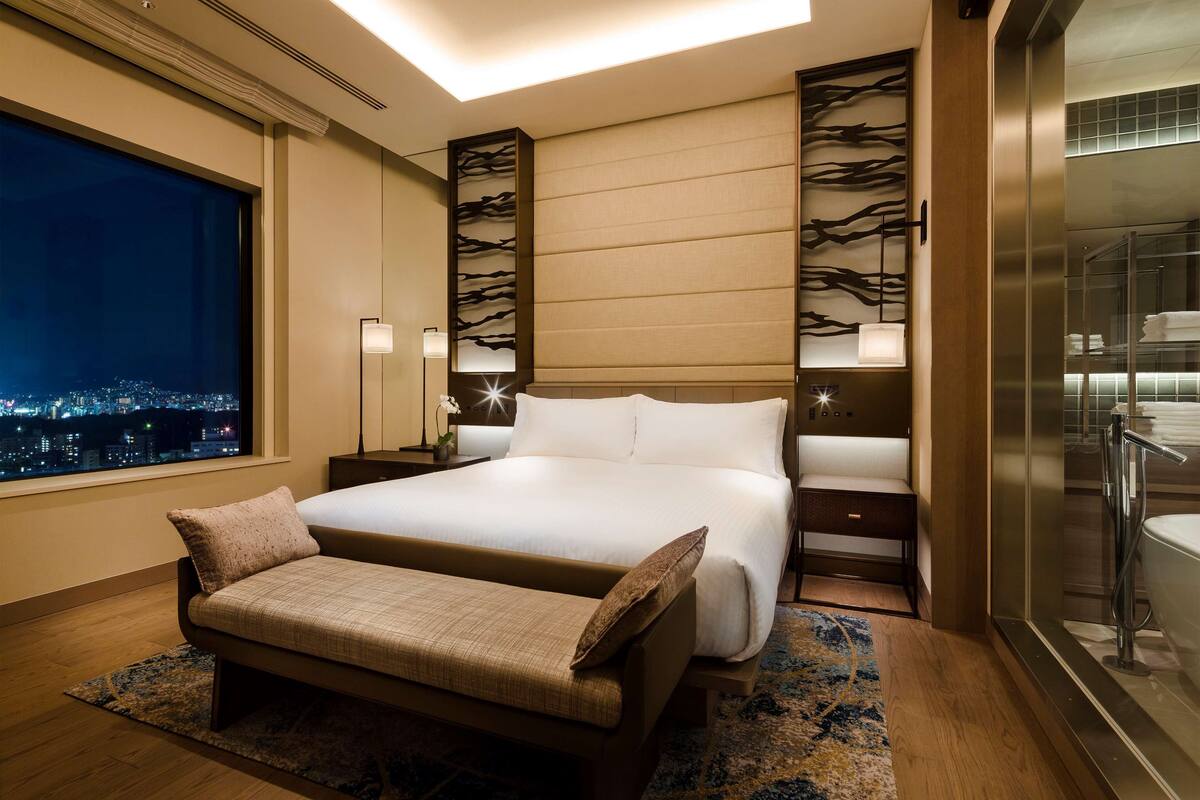 Habitación de hotel moderna con una cama grande, mesitas de noche, un banco, vistas a la ciudad y un baño con ducha de vidrio.