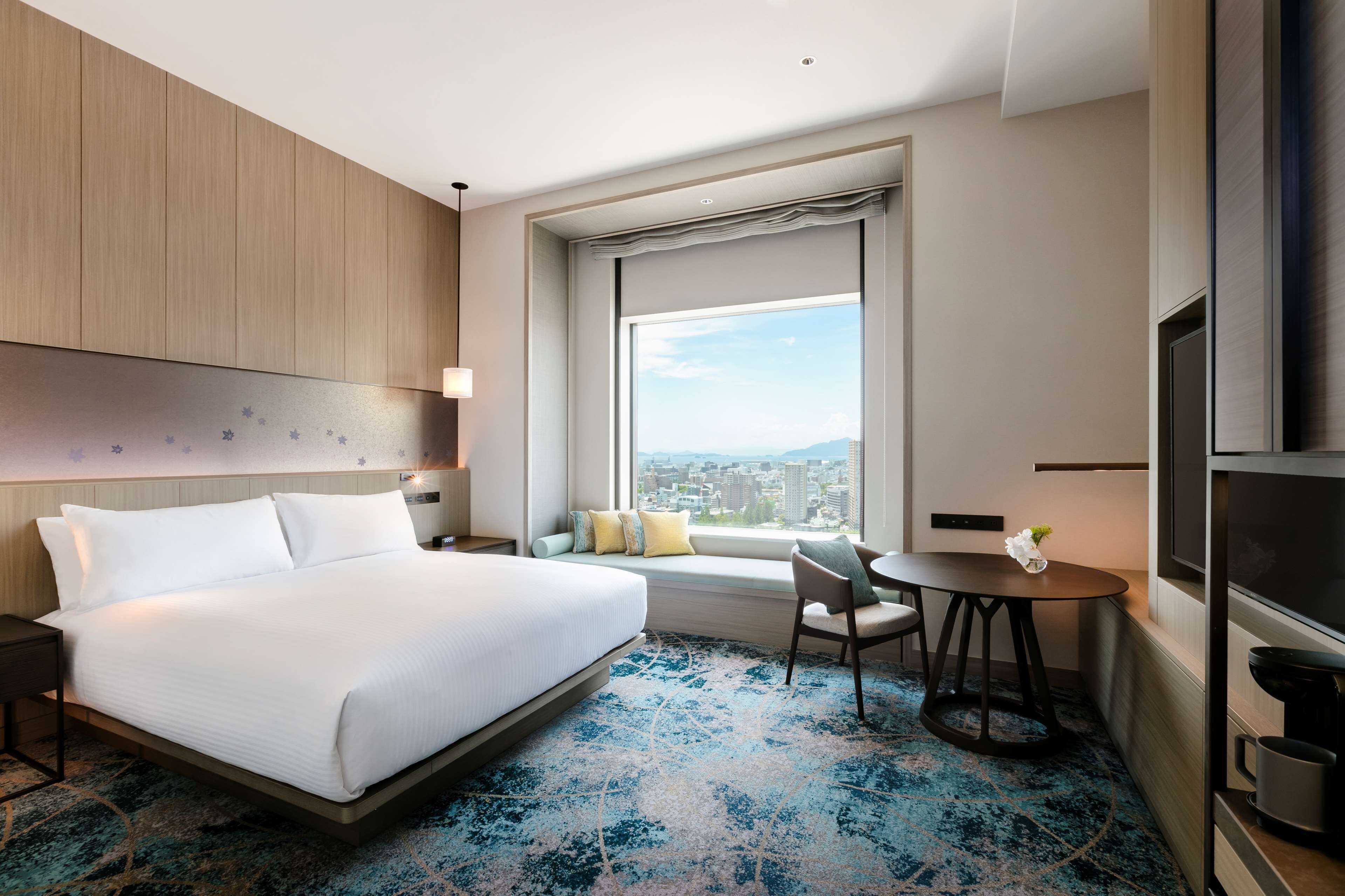 Habitación de hotel con una cama grande, una mesita con un florero, un ventanal con vista a la ciudad y un televisor a la derecha.