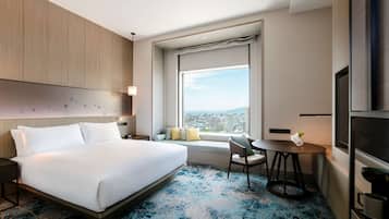Habitación de hotel con una cama grande, una mesita con un florero, un ventanal con vista a la ciudad y un televisor a la derecha.