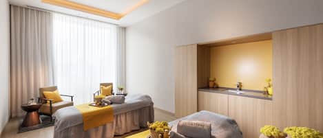 A modern hotel room with two beds, a sitting area with a chair and table, a vanity with a mirror, and a large window with curtains.