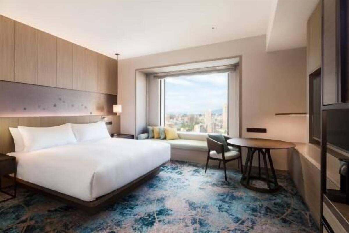 Habitación de hotel con cama, escritorio, silla, ventana y vista a la ciudad, y televisor.
