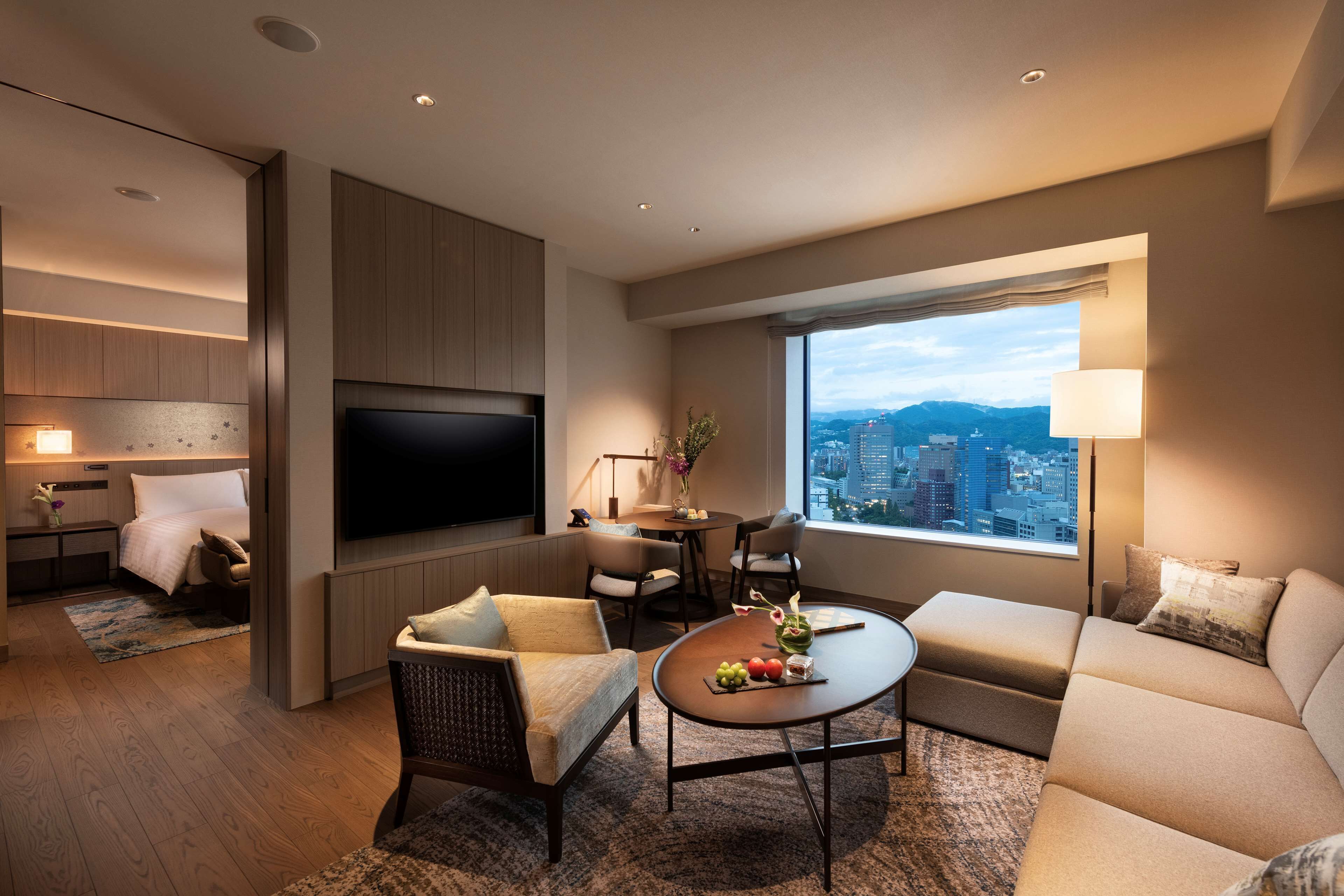 Ein modernes Hotelzimmer mit Flachbildfernseher, einer Sitzecke, einem Essbereich und einem großen Fenster mit Blick auf die Stadt.