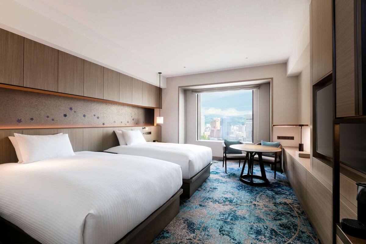 Habitación de hotel con dos camas, una pequeña mesa de comedor, una silla, un ventanal con vistas a la ciudad y un televisor de pantalla plana.