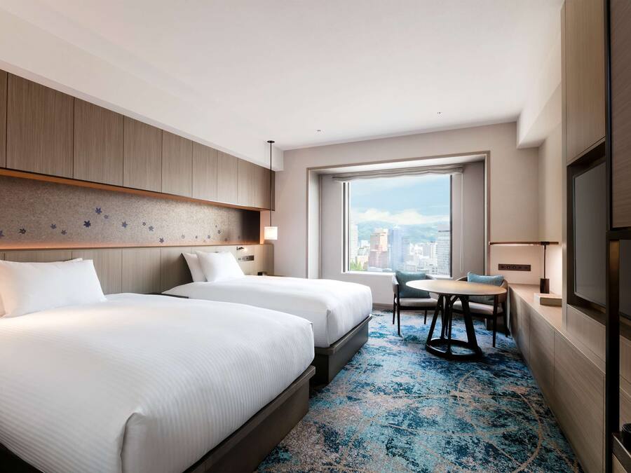 Une chambre d’hôtel avec deux lits, une petite table à manger, une chaise, une grande fenêtre donnant sur la ville et un téléviseur à écran plat.