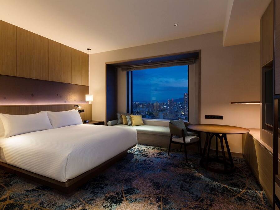 A modern hotel room with a large bed, a seating area with a view of the city at night, and a small table.