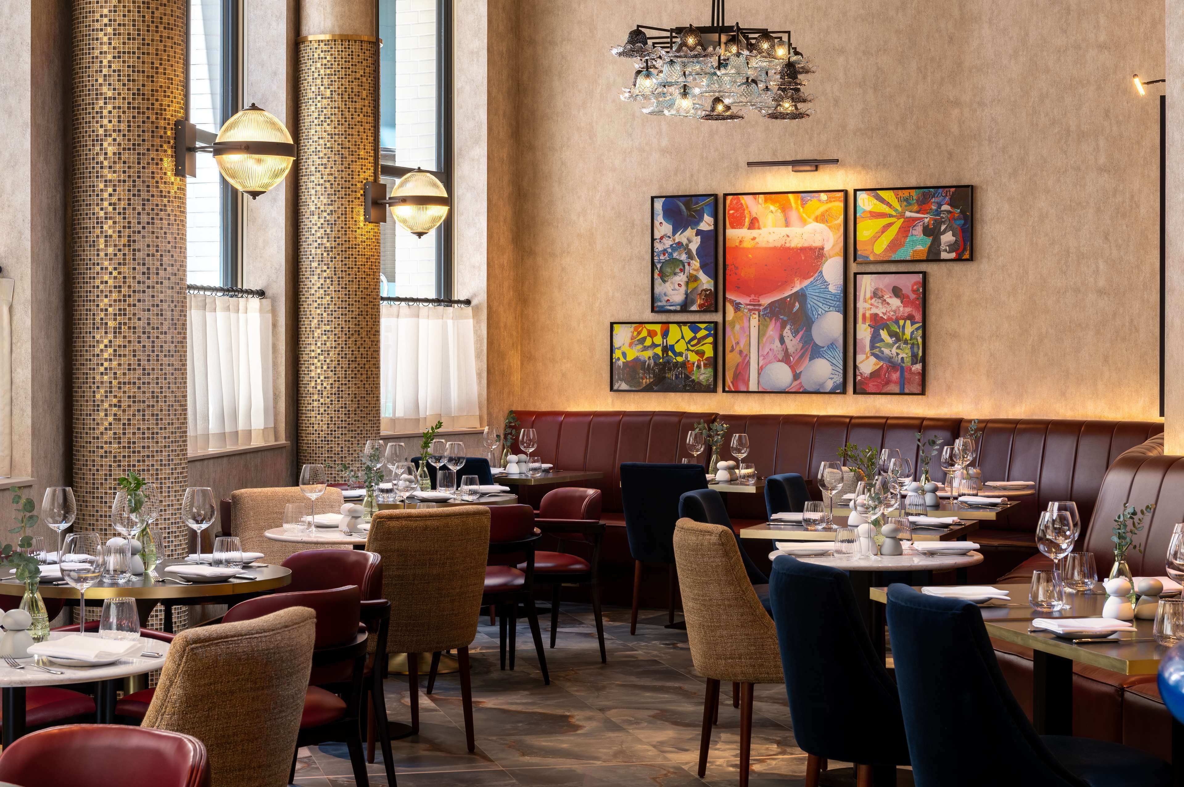 Un restaurante con mesas redondas, sillas rojas y azules, y una pared decorada con obras de arte coloridas.