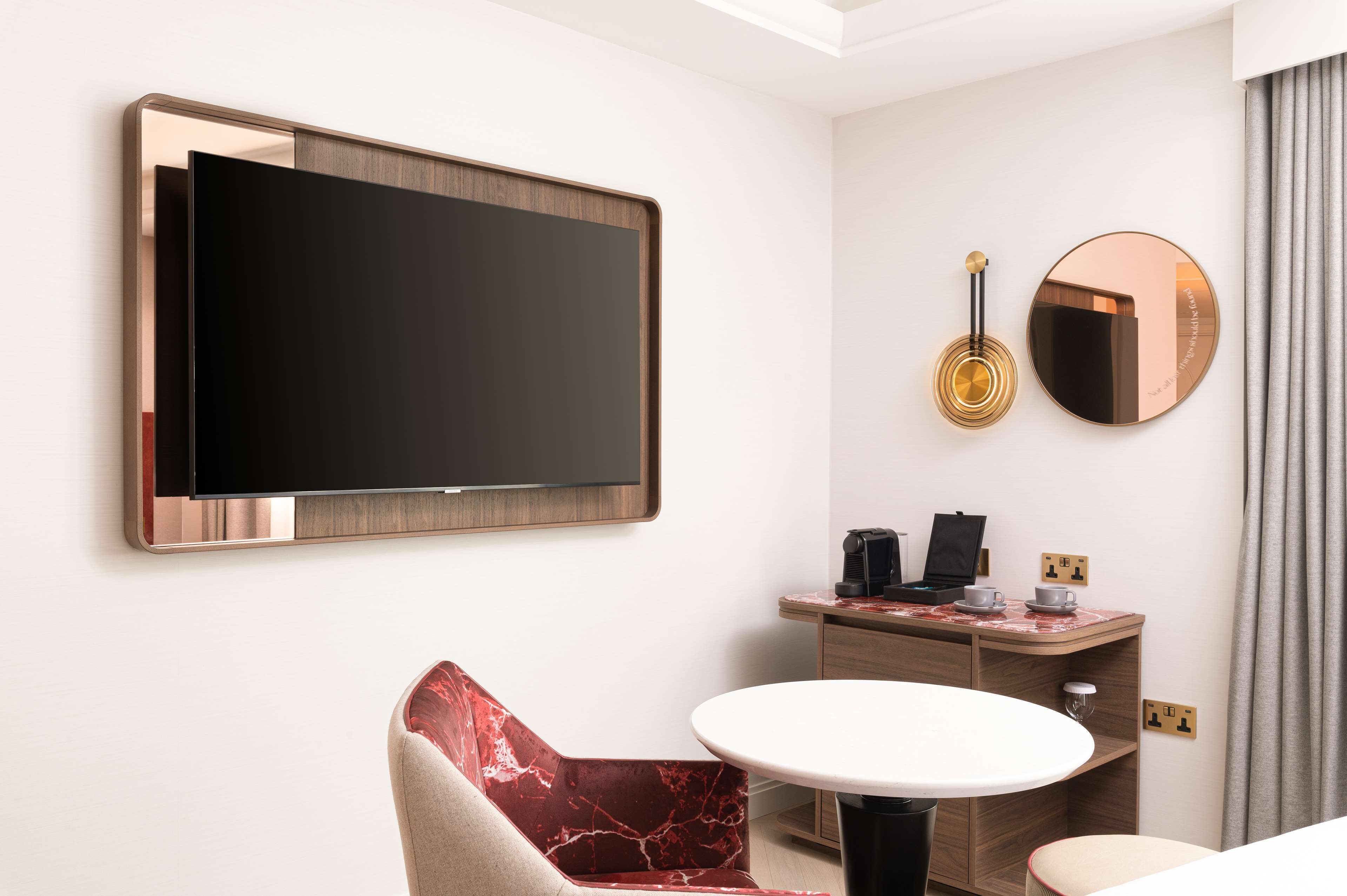 A hotel room with a large flat-screen TV mounted on the wall, a round mirror, a small desk with a coffee maker, and a red and beige chair.