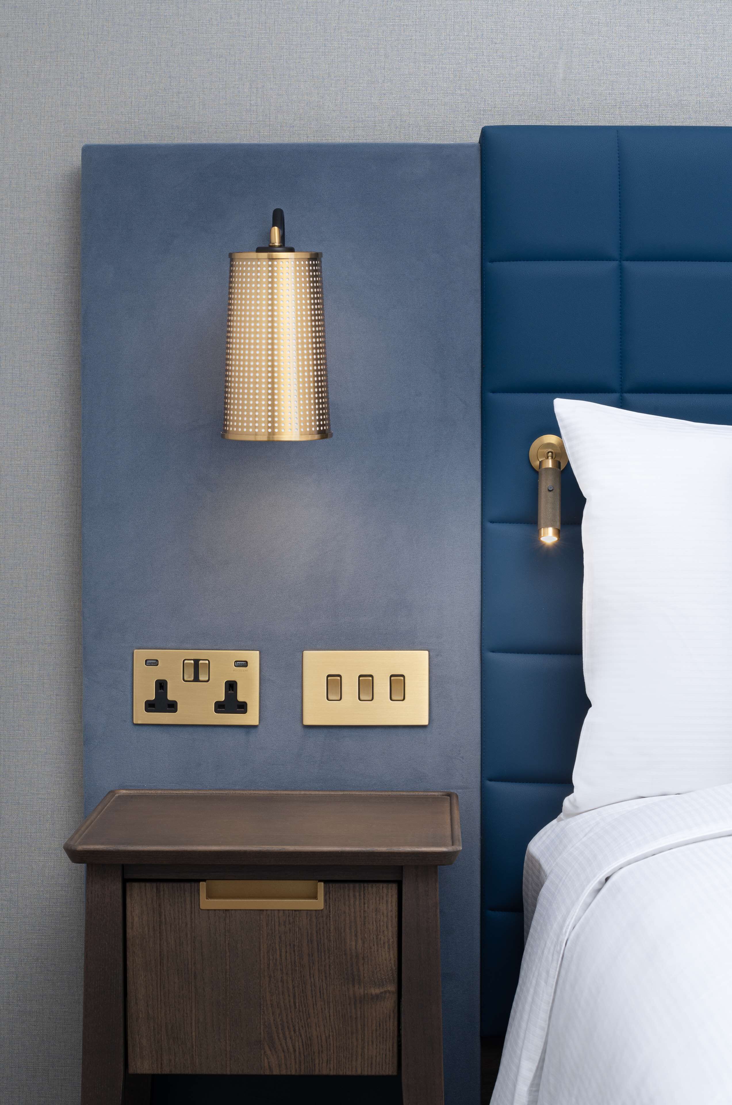 Ein Schlafzimmer mit blauem Kopfteil, einer Nachttischlampe, einem Nachttisch und einem Bett mit weißer Bettwäsche.