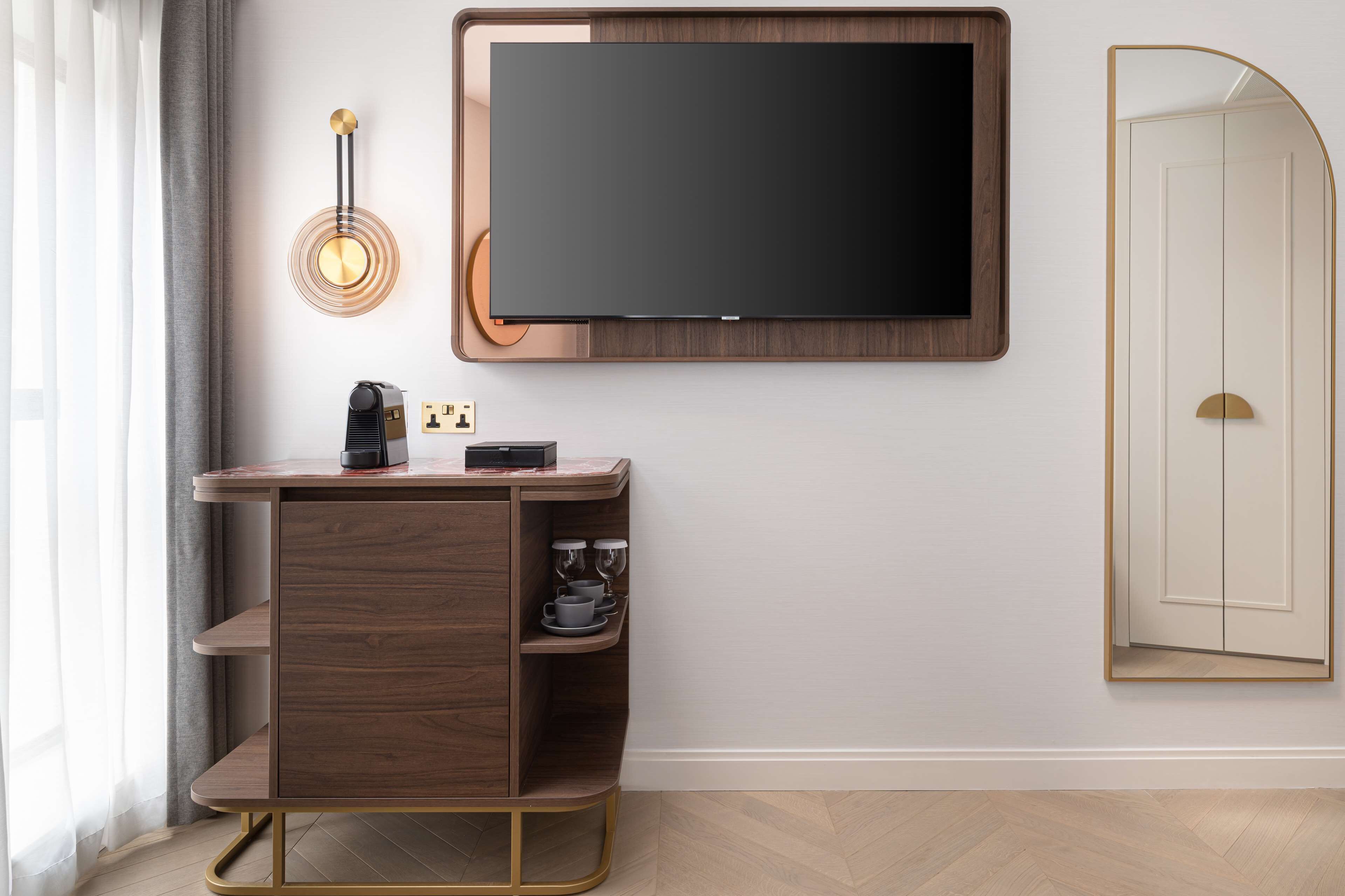 A modern hotel room with a flat-screen TV mounted on the wall, a wooden console table with a coffee machine, and a mirror with a gold frame.